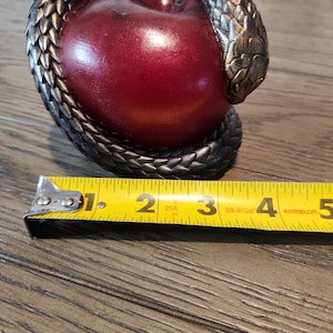 May include: A decorative sculpture featuring a red apple encircled by a detailed, metallic snake. The snake's scales are textured, and the apple has a glossy finish. The sculpture is approximately 4 inches long.