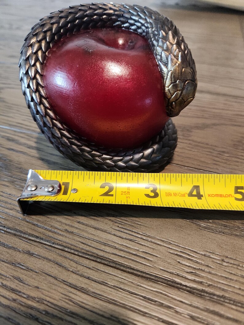 May include: A decorative sculpture featuring a red apple encircled by a detailed, metallic snake. The snake's scales are textured, and the apple has a glossy finish. The sculpture is approximately 4 inches long.