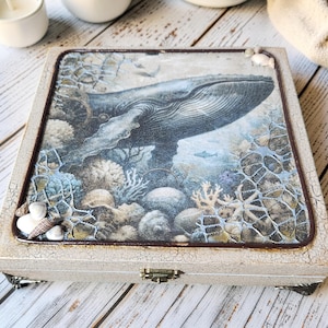 May include: A decorative wooden box with a whale illustration. The box features a distressed white finish, a hinged lid, and a detailed image of a whale swimming in an underwater scene with coral and seashells. The box is approximately 6 inches square.
