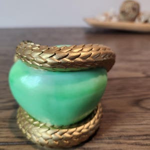 May include: A green heart-shaped object is encircled by a gold-colored, textured band. The band appears to be made of overlapping scales. The object sits on a wooden surface.