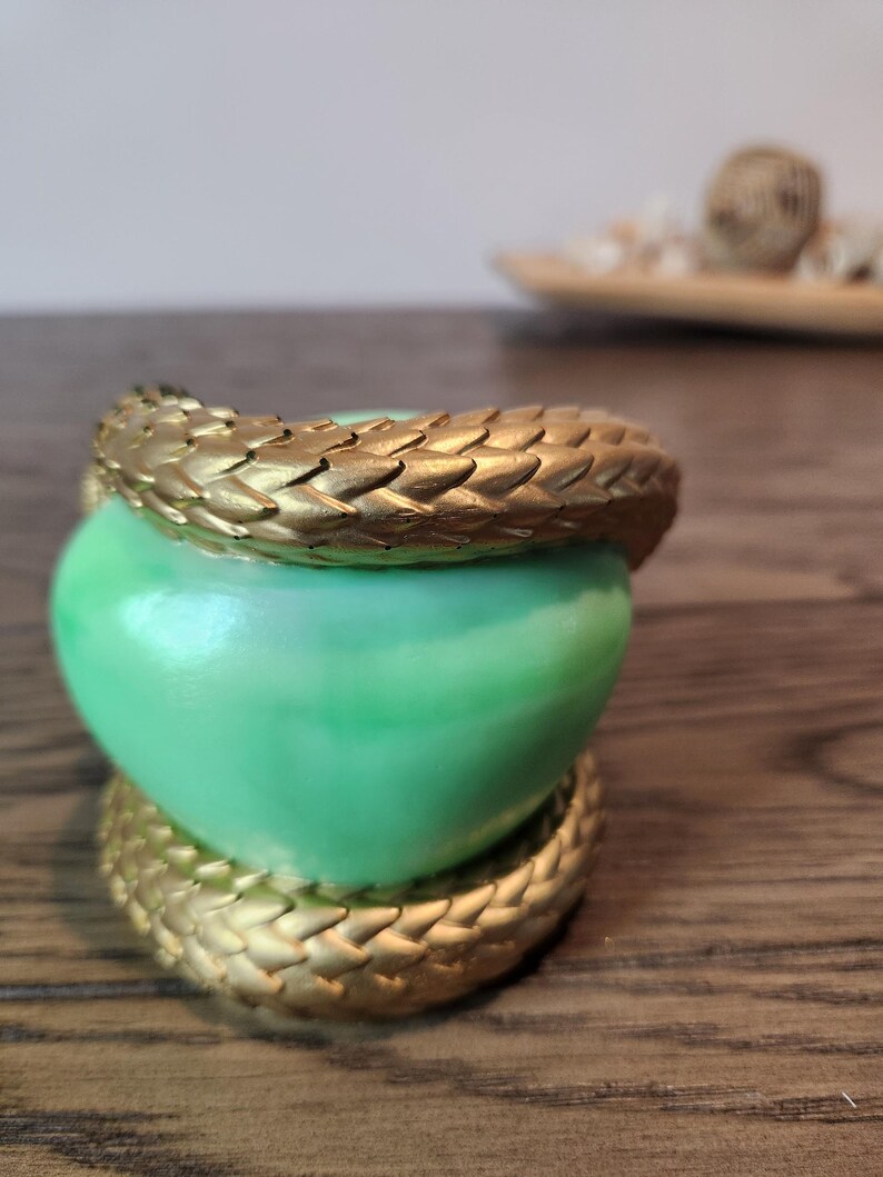 May include: A green heart-shaped object is encircled by a gold-colored, textured band. The band appears to be made of overlapping scales. The object sits on a wooden surface.