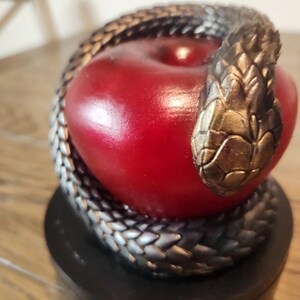 May include: A decorative sculpture featuring a red apple encircled by a silver and gold-toned snake. The snake's head rests atop the apple. The sculpture sits on a black base, creating a striking contrast.