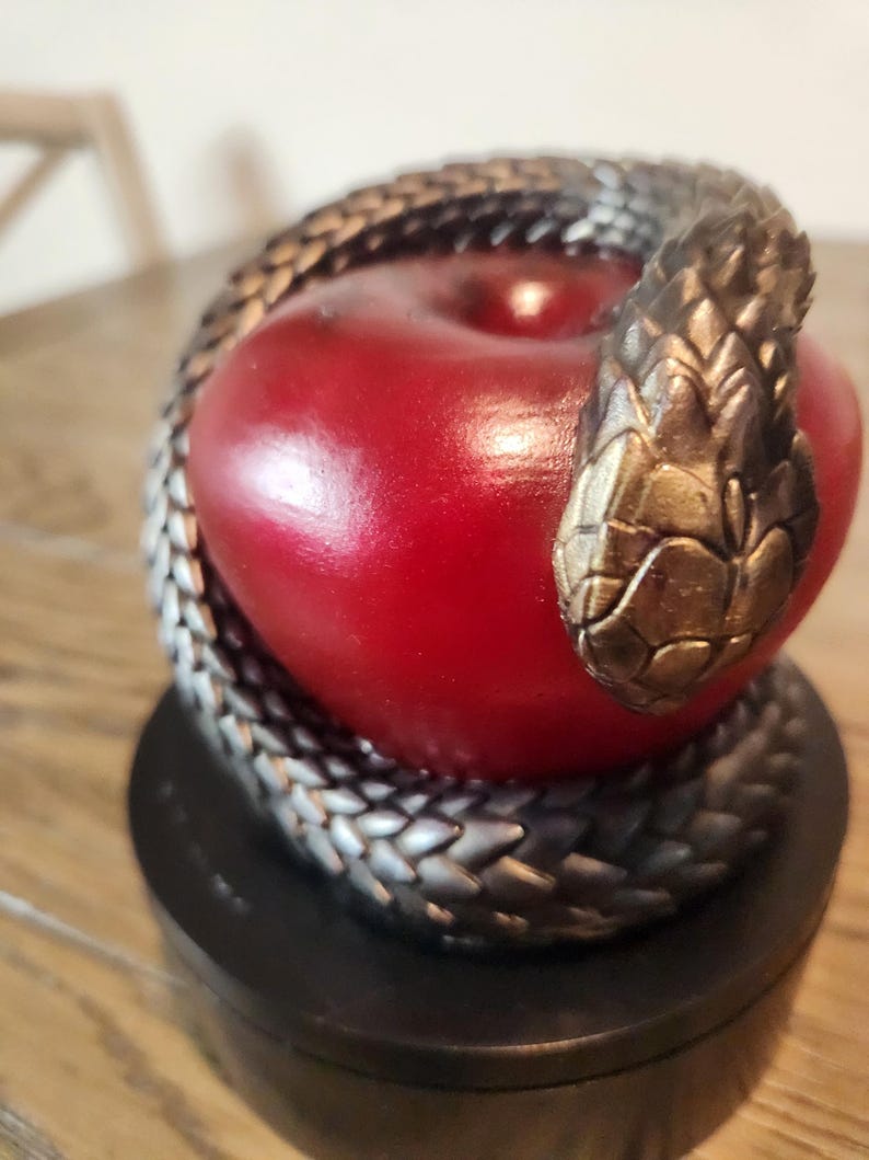 May include: A decorative sculpture featuring a red apple encircled by a silver and gold-toned snake. The snake's head rests atop the apple. The sculpture sits on a black base, creating a striking contrast.