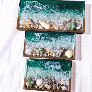 May include: Three rectangular resin trays with a beach scene design. Each tray features a dark green ocean, white foamy waves, and a sandy shore with seashells, pebbles, and starfish. The trays have a brown border.