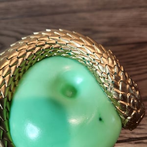 May include: A gold-colored, textured snake encircles a light green, rounded object. The snake's scales are detailed, and the object has a smooth surface with a small indentation. The background is a wooden surface.