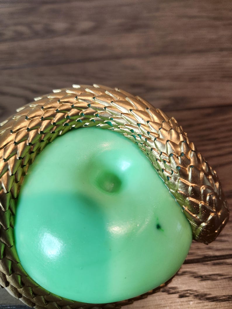 May include: A gold-colored, textured snake encircles a light green, rounded object. The snake's scales are detailed, and the object has a smooth surface with a small indentation. The background is a wooden surface.