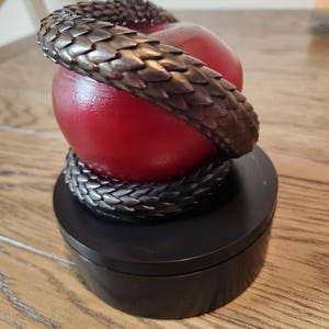 May include: A decorative sculpture featuring a red apple encircled by a bronze-colored snake. The snake's scales are detailed, and the apple rests on a black, round base. The sculpture is a unique home decor item.