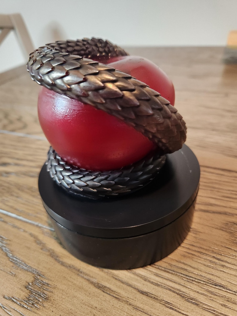 May include: A decorative sculpture featuring a red apple encircled by a bronze-colored snake. The snake's scales are detailed, and the apple rests on a black, round base. The sculpture is a unique home decor item.