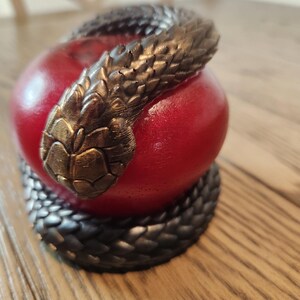 May include: A red apple sculpture with a dark bronze snake coiled around it. The snake's head is positioned at the top of the apple. The sculpture is on a wooden surface.