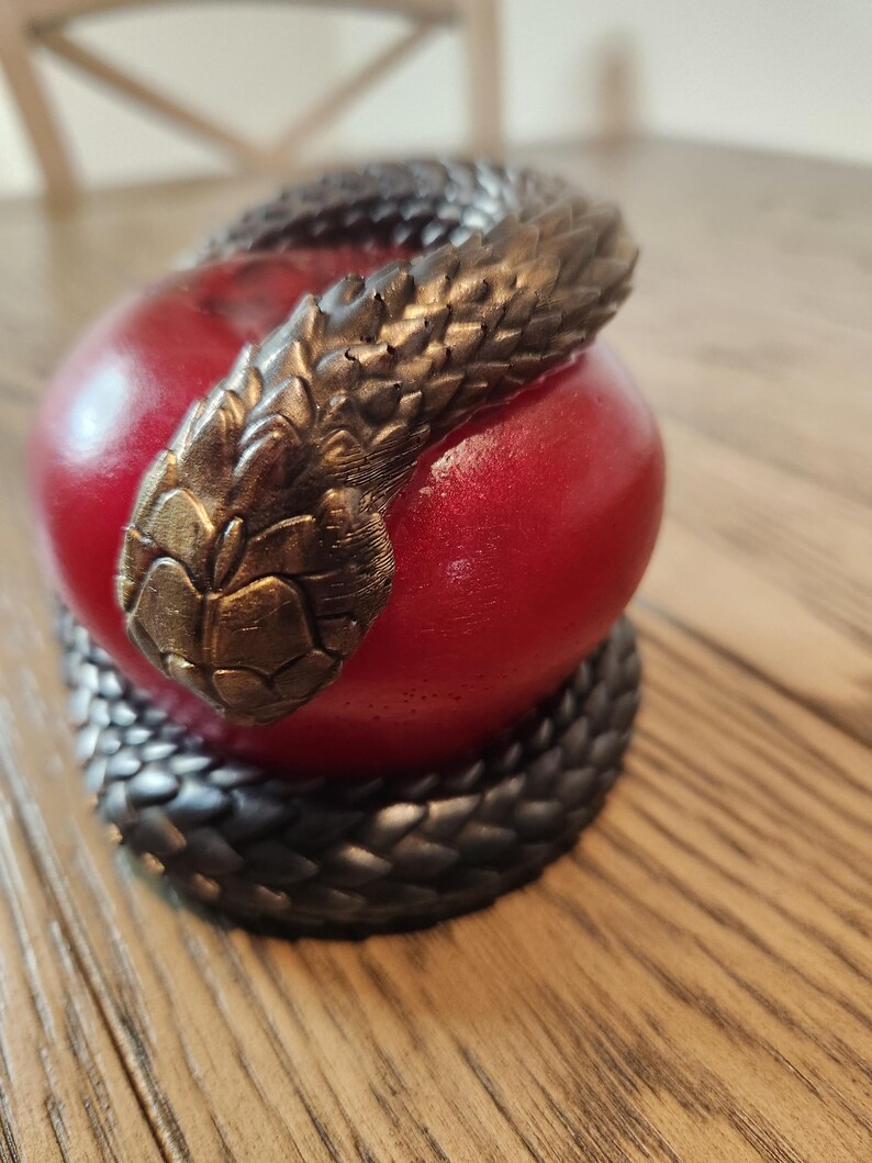 May include: A red apple sculpture with a dark bronze snake coiled around it. The snake's head is positioned at the top of the apple. The sculpture is on a wooden surface.