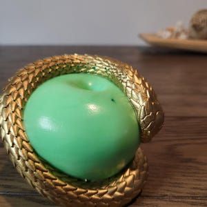 May include: A gold-colored snake coiled around a green apple. The snake has detailed scales and is wrapped around the apple, creating a decorative piece. The apple is a vibrant green color.