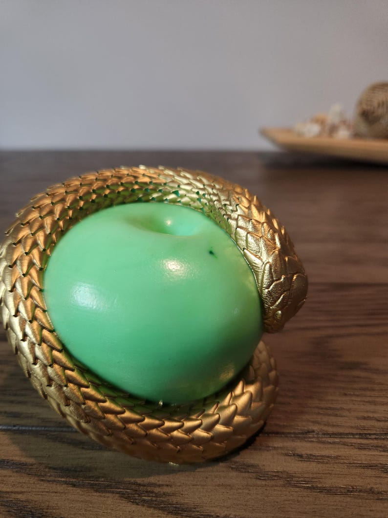 May include: A gold-colored snake coiled around a green apple. The snake has detailed scales and is wrapped around the apple, creating a decorative piece. The apple is a vibrant green color.