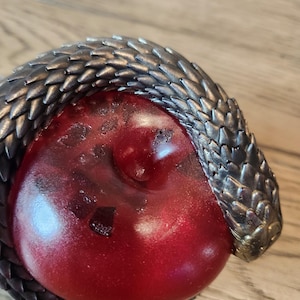 May include: A detailed close-up of a decorative object featuring a red, rounded form encircled by a metallic, textured snake. The snake's scales are a dark, gunmetal color, contrasting with the glossy red center. The object is set against a wooden surface.