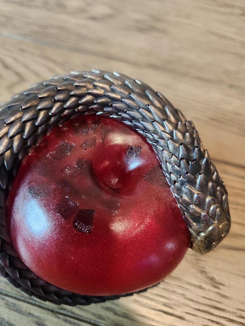 May include: A detailed close-up of a decorative object featuring a red, rounded form encircled by a metallic, textured snake. The snake's scales are a dark, gunmetal color, contrasting with the glossy red center. The object is set against a wooden surface.