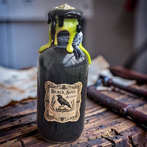 May include: A glass bottle filled with a dark, shimmering liquid, topped with dripping black and neon green wax. A rectangular label features an ornate border, a black crow illustration, skulls, and the text Black Soul Elixir.