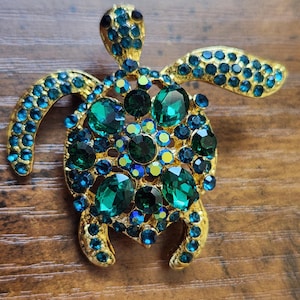 May include: A gold-tone turtle brooch featuring a shell encrusted with large emerald green faceted crystals and smaller blue rhinestones. The head and flippers are also decorated with blue rhinestones, and the eyes are set with small black stones.
