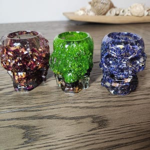May include: Three resin skull-shaped candle holders. One is filled with red and gold, the second with green, and the third with blue and white. The candle holders are on a wooden surface.