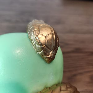 May include: A gold-colored snake head adorns a light green, rounded object. The snake's head is detailed with scales and sits atop a textured, gold-colored base. The design suggests a decorative item or sculpture.