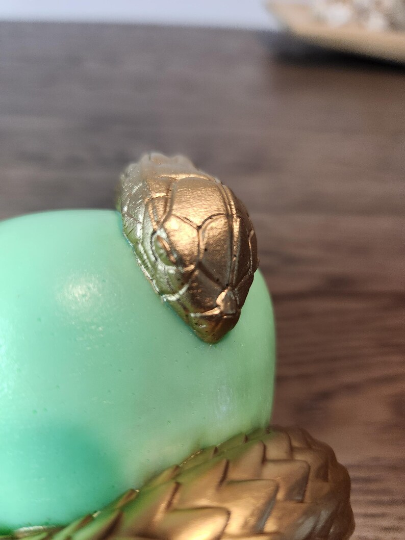 May include: A gold-colored snake head adorns a light green, rounded object. The snake's head is detailed with scales and sits atop a textured, gold-colored base. The design suggests a decorative item or sculpture.
