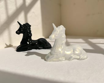 May include: Two small unicorn figurines. One is glossy black, the other is iridescent white. Both are in a resting pose, with their front legs tucked beneath them. The unicorns have a horn and flowing mane.
