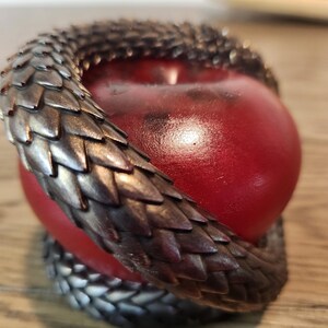 May include: A red apple encircled by a metallic, scale-like sculpture. The sculpture wraps around the apple, creating a decorative and unique design. The apple has a glossy finish, and the sculpture has a dark, textured appearance.