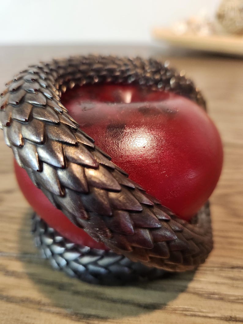 May include: A red apple encircled by a metallic, scale-like sculpture. The sculpture wraps around the apple, creating a decorative and unique design. The apple has a glossy finish, and the sculpture has a dark, textured appearance.