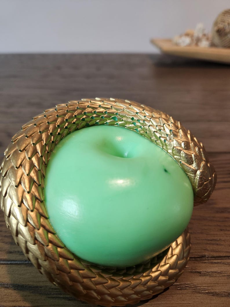 May include: A green apple encircled by a gold-colored, textured snake. The snake's scales are detailed, and the apple has a smooth surface. The composition is set against a blurred wooden background.