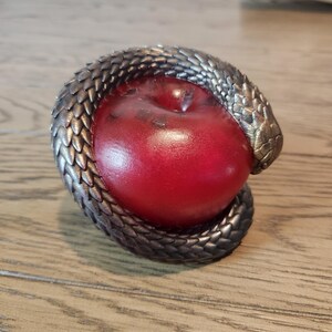 May include: A red apple encircled by a metallic, snake-like sculpture. The snake's scales are detailed with a bronze and silver finish. The apple has a glossy surface, creating a striking contrast with the textured snake.