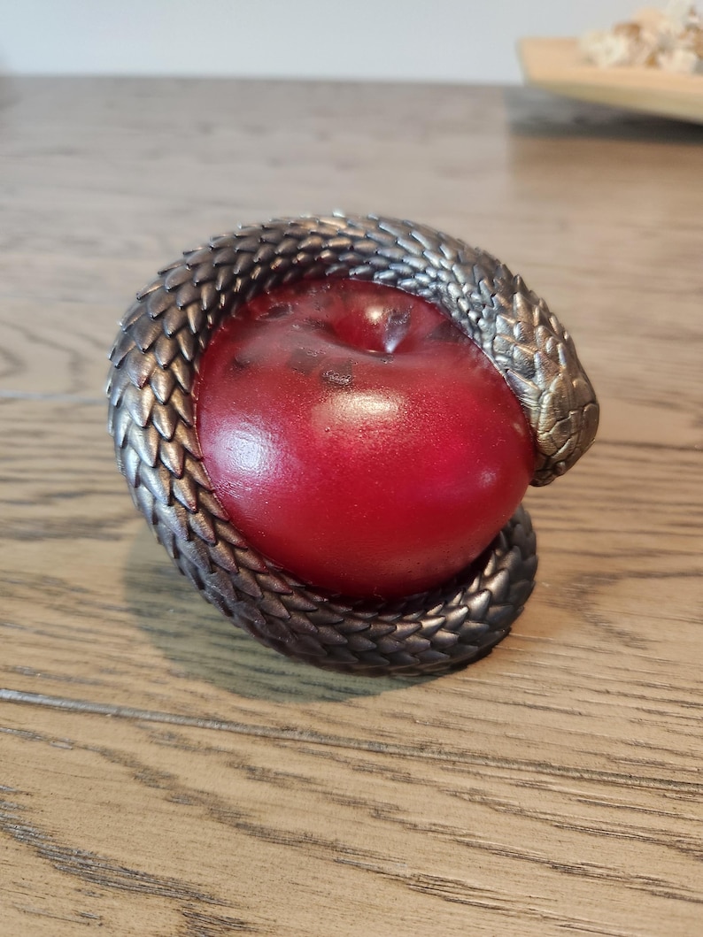 May include: A red apple encircled by a metallic, snake-like sculpture. The snake's scales are detailed with a bronze and silver finish. The apple has a glossy surface, creating a striking contrast with the textured snake.