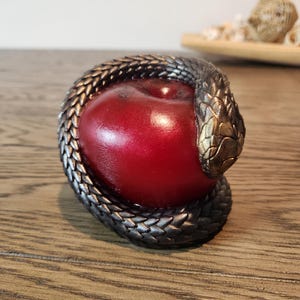 May include: A decorative sculpture featuring a vibrant red apple encircled by a detailed, metallic snake. The snake's scales are intricately designed, with a bronze-colored head. The sculpture is set on a wooden surface.