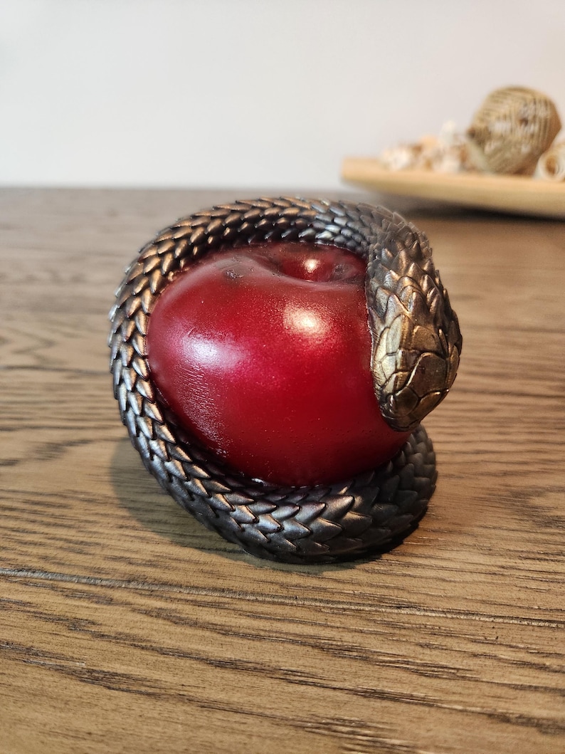 May include: A decorative sculpture featuring a vibrant red apple encircled by a detailed, metallic snake. The snake's scales are intricately designed, with a bronze-colored head. The sculpture is set on a wooden surface.