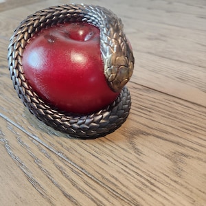 May include: A decorative sculpture featuring a red apple encircled by a detailed, metallic snake. The snake's head is bronze-colored, and the body is a darker, textured metal. The apple is smooth and glossy.