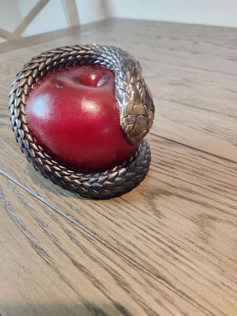 May include: A decorative sculpture featuring a red apple encircled by a detailed, metallic snake. The snake's head is bronze-colored, and the body is a darker, textured metal. The apple is smooth and glossy.