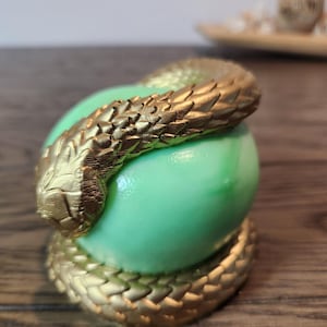 May include: A gold-colored snake coiled around a light green sphere. The snake has detailed scales and is wrapped around the sphere, with the tail coiled at the base. The background is blurred, with a wooden surface.