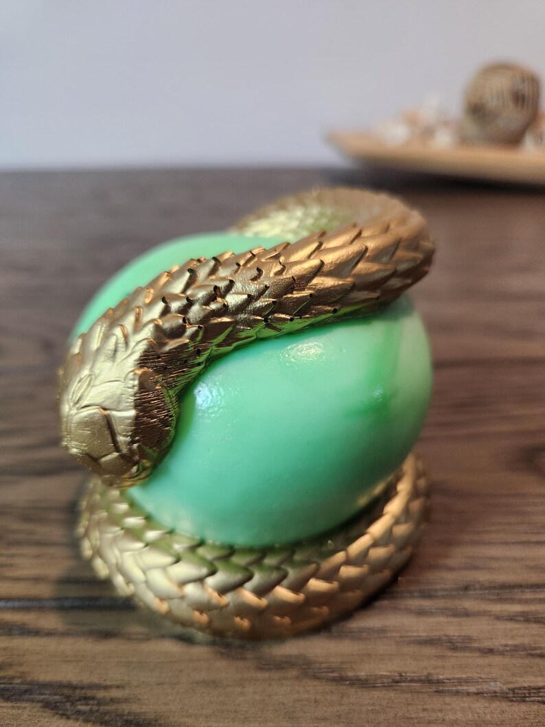 May include: A gold-colored snake coiled around a light green sphere. The snake has detailed scales and is wrapped around the sphere, with the tail coiled at the base. The background is blurred, with a wooden surface.