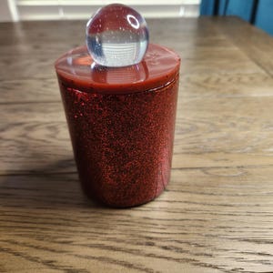 May include: A cylindrical container with a red glitter finish. The container is topped with a solid red lid and a clear glass orb. The container is sitting on a wooden surface.