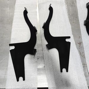 Op de afbeelding: Twee zwarte metalen beugels met een gebogen, langwerpig ontwerp. Elke beugel heeft een montageplaat aan de bovenkant en een gekerfde basis. De beugels worden op een wit oppervlak weergegeven.