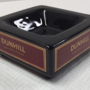 May include: A square black ceramic ashtray featuring a deep basin and a maroon band on the sides. The text "DUNHILL" is printed in gold lettering above the words "London-Paris-New York" on the maroon sections, framed by thin gold border lines.