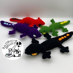 May include: A collection of handmade crochet lizard toys in various colors: red, purple, green, and black. Each lizard features small details like bead accents and contrasting eye colors. The toys are arranged on a white surface.