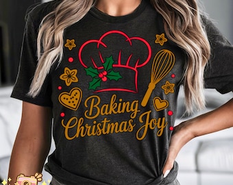 May include: Dark gray t-shirt with a festive design. The graphic features a red chef's hat, a whisk, and the words "Baking Christmas Joy" in gold. Additional elements include holly, stars, hearts, and gingerbread cookies.