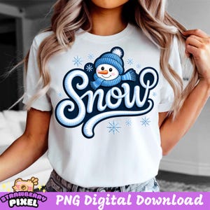 Peut inclure: T-shirt blanc avec un motif sur le thème de l'hiver. Le design présente le mot "Snow" dans une police bleue stylisée, accompagné de flocons de neige et d'un bonhomme de neige portant un bonnet et une écharpe bleus. Le t-shirt est de style décontracté à manches courtes.