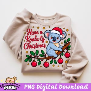 Może przedstawiać: Beżowa bluza z nadrukiem koali w czapce Świętego Mikołaja, siedzącego na gałęzi z czerwonymi bombkami. Tekst brzmi "Have a Koala-ty Christmas" w czerwonym skrypcie ze złotymi gwiazdami.