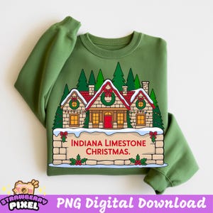 May include: A green sweatshirt with a Christmas-themed design. The design shows a house with a red roof, wreaths, and fairy lights, with evergreen trees in the background. The text "INDIANA LIMESTONE CHRISTMAS" is displayed on a stone sign.