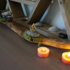 May include: A wooden branch with moss and a small golden statue on a rock. Two lit candles are on a wooden surface.