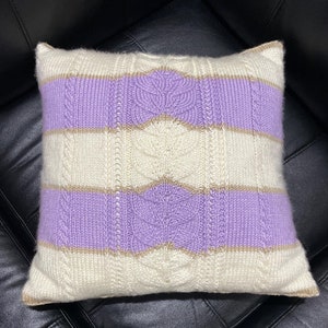 May include: A knitted throw pillow with a white background and purple and brown stripes. The pillow has a cable knit pattern.
