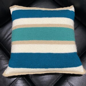 May include: A striped knitted pillow with a beige, white, teal, and dark blue color scheme. The pillow has a beige trim around the edges.