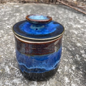Op de afbeelding: Een blauwe en bruine keramische pot met een deksel. De pot heeft een donkerblauwe glazuur met bruine en witte accenten. Het deksel is donkerblauw met een bruine rand.