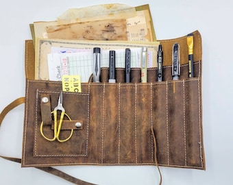 May include: A brown leather roll-up case with multiple slots for holding pens and pencils. The case has a strap and a small pocket for a pair of scissors. The case is filled with various pens and pencils, including a black pen with the word "BLACK" on it.