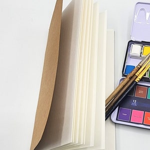 Può includere: Un album da disegno marrone con pagine bianche è aperto per rivelare la carta bianca all'interno. Tre pennelli sono appoggiati sull'album da disegno. Un set di colori ad acquerello con 12 colori è visibile sullo sfondo.