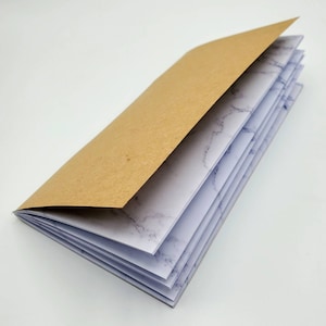 May include: A brown paper covered notebook with white pages featuring a marble pattern. The notebook is open to reveal the pages.